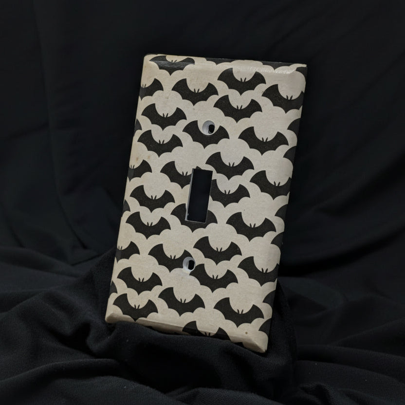 A single light switch plate cover featuring a repeating black bat pattern on a warm cream background, displayed against a draped black fabric backdrop.