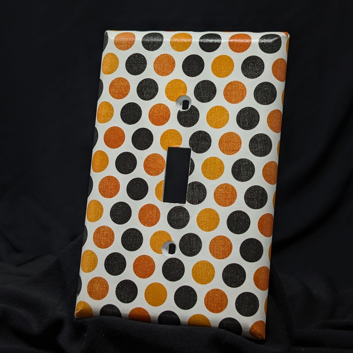 A single-toggle light switch plate cover featuring a pattern of orange, black, and dark brown polka dots on a white background, displayed against a black fabric backdrop.