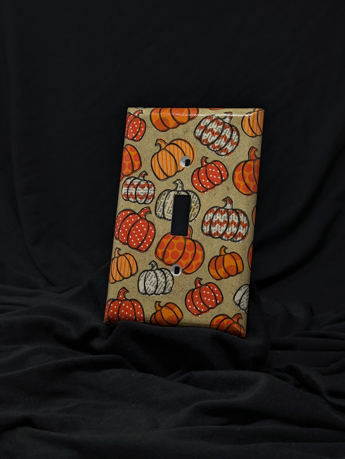 A single light switch cover plate featuring a fall harvest pumpkin pattern, displayed against a draped black fabric background.