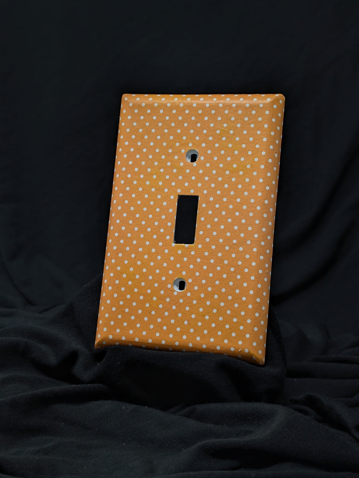 An orange single-toggle light switch plate cover decorated with a white polka dot pattern, displayed on a draped black fabric background.